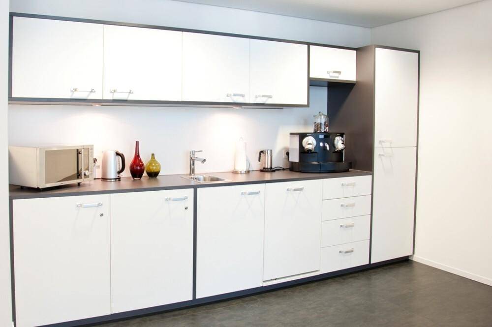 Moderne Küchenzeile mit weißen Schränken, Mikrowelle, Kaffeemaschine und Spüle für den Bürogebrauch.