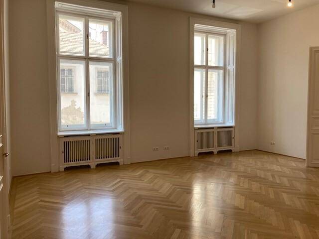Heller Raum mit Fischgrätparkett und zwei großen Fenstern, die den Blick nach draußen freigeben und Licht hereinlassen.
