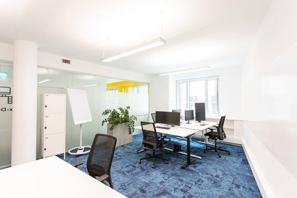 Modernes Büro mit blauer Teppichboden, weißen Schreibtischen und einem Whiteboard.