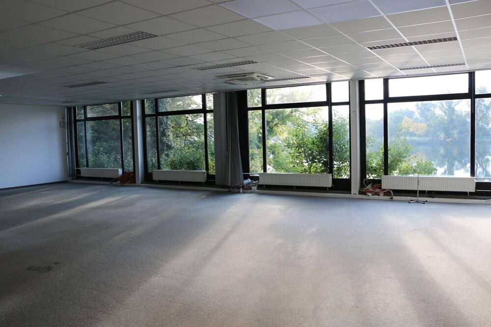 Großer, leerer Büroraum mit Teppichboden und weitem Blick auf Bäume und Wasser durch die Fensterfront.