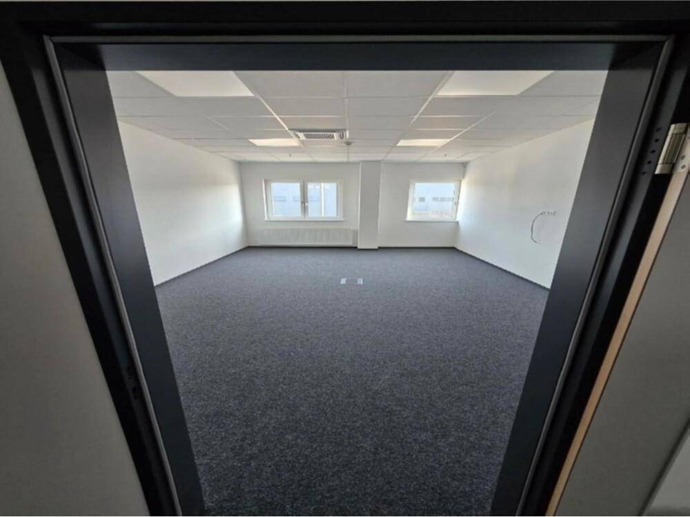 Blick durch eine offene Tür in einen leeren Büroraum mit grauem Teppichboden und hellen Wänden.