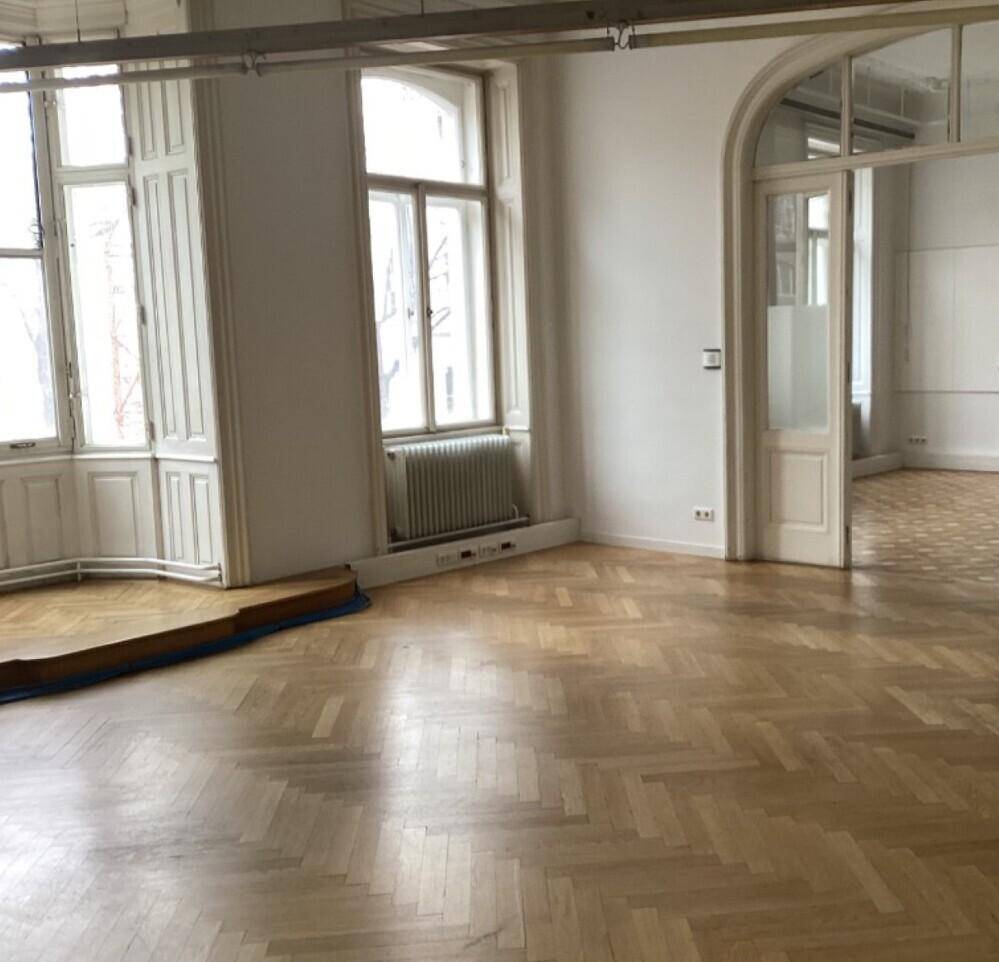 Großes, helles Zimmer mit Fischgrätparkett, hohen Fenstern und einer Nische.