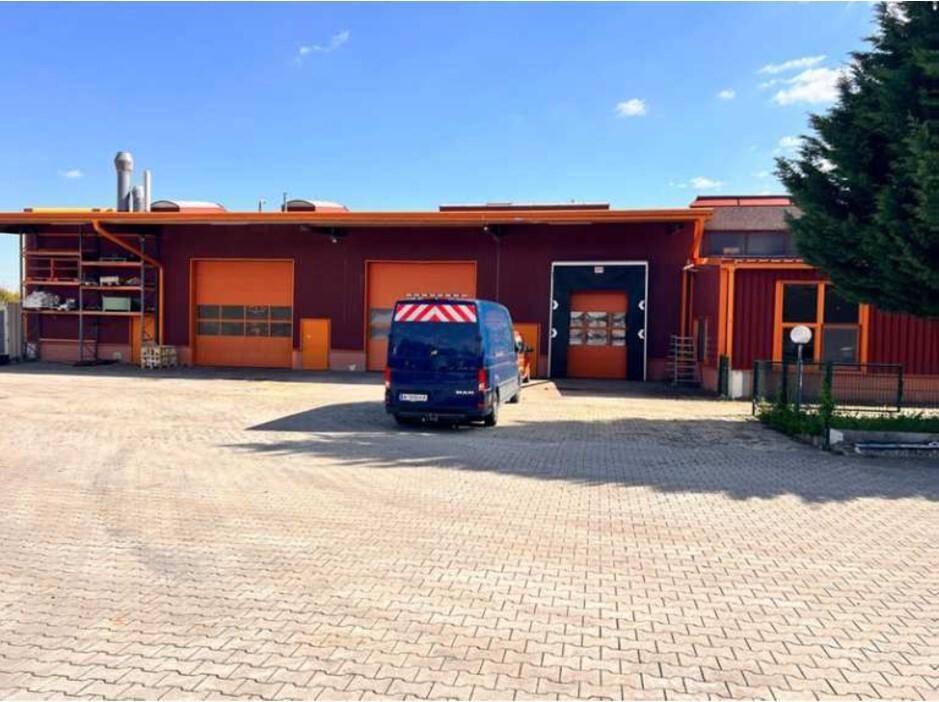 Industriegebäude mit roter Fassade, mehreren großen orangefarbenen Rolltoren und einem gepflasterten Vorplatz mit Lieferwagen.