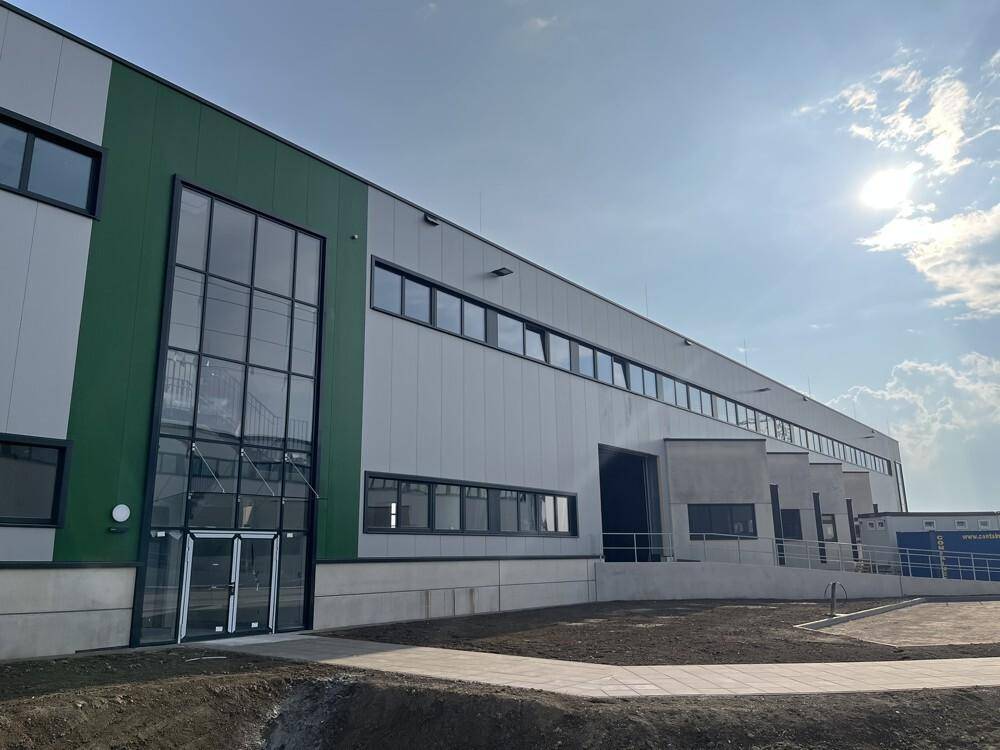 Die moderne Außenansicht des Industriegebäudes mit Glasportal, Laderampen und der grau-grünen Fassade bei Sonnenschein.