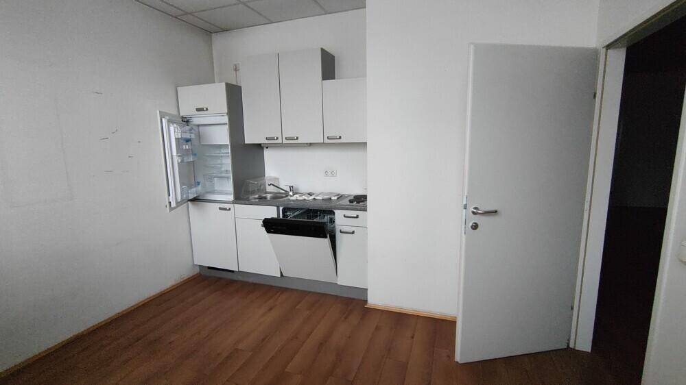 Kleine Einbauküche mit weißen Schränken, Spülmaschine und Kühlschrank in einem Büroraum.
