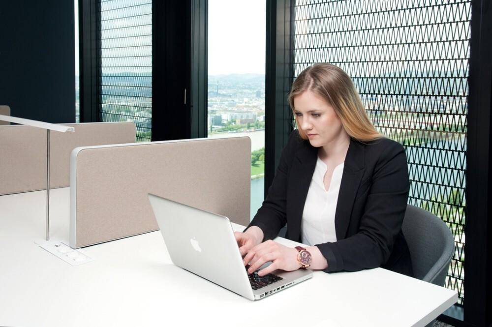 Eine Frau arbeitet an einem Laptop an einem Schreibtisch mit Trennwänden und Fensterblick.