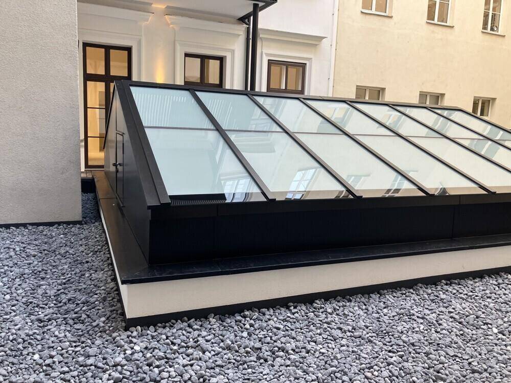 Modernes Glasdach über einem Innenhof, umgeben von Kies und den Wänden des Gebäudes.