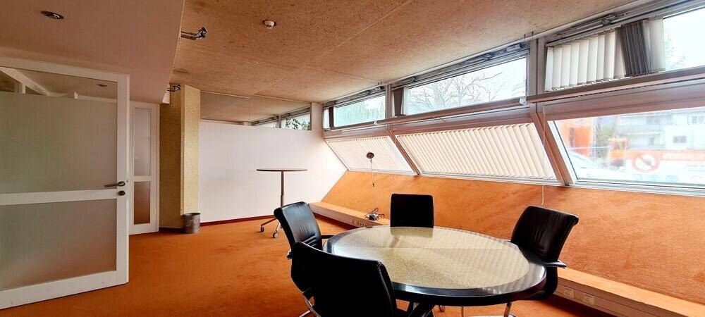 Besprechungsraum mit rundem Tisch, Stühlen und Fenstern mit Jalousien auf orangefarbenem Teppichboden.