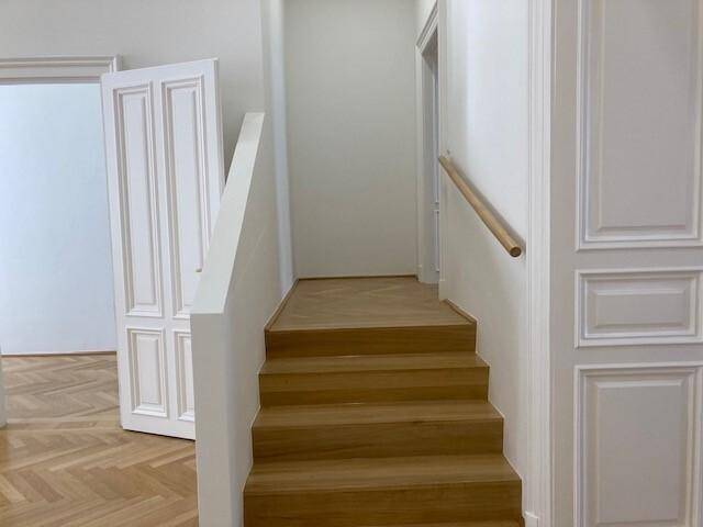 Holztreppe mit schlichtem weißen Geländer und Fischgrätparkett im unteren Bereich, umgeben von hellen Wänden.