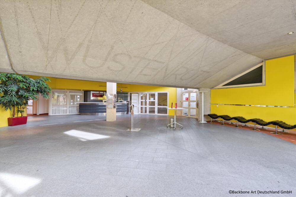 Geräumige Lobby mit leuchtend gelben Wänden, grauem Fliesenboden und einem modernen Empfangsbereich.