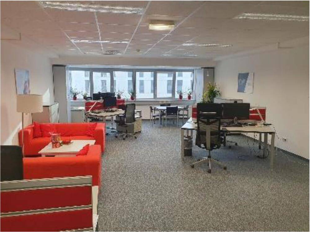 Heller Büroraum mit roten Sofas, mehreren Arbeitsplätzen und großen Fenstern, die viel Tageslicht hereinlassen.