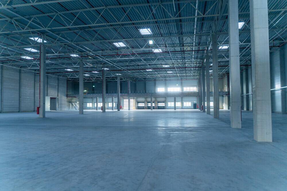 Geräumige, leere Lagerhalle mit hoher Decke, vielen Stützen und Oberlichtern für natürliches Licht.