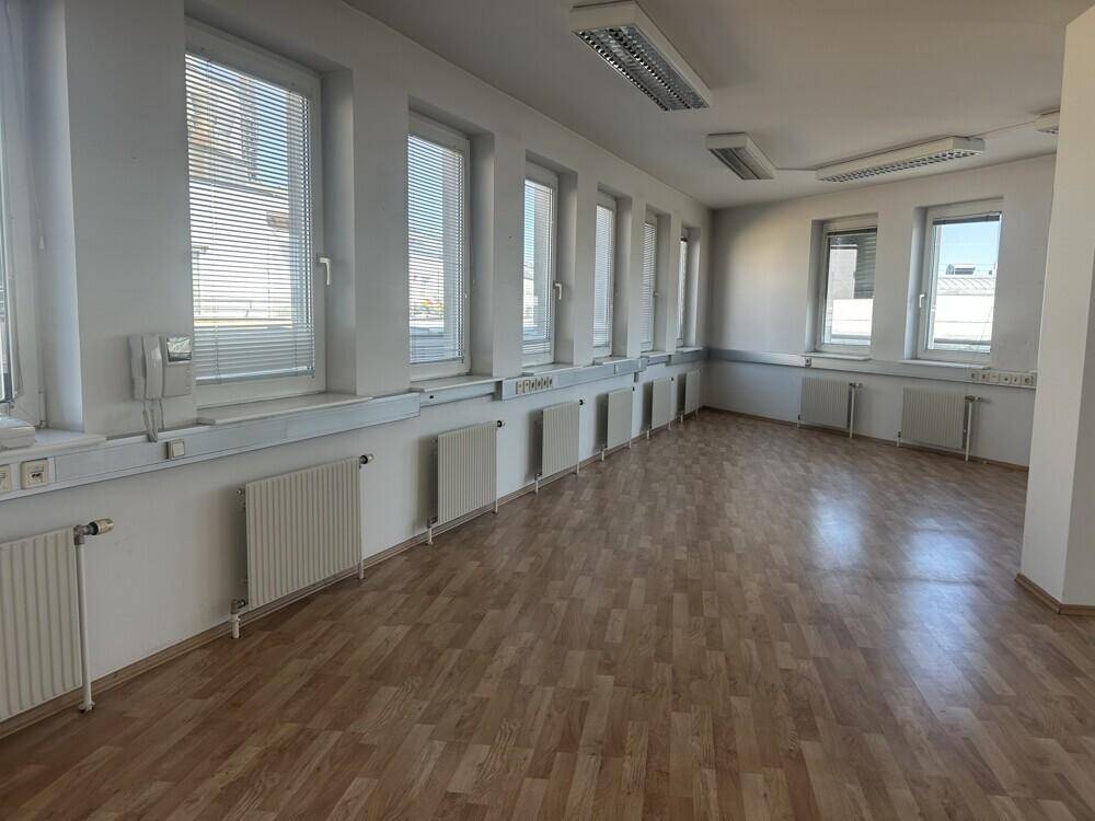 Langer Büroraum mit vielen Fenstern, Jalousien, Heizkörpern und hellem Laminatboden.