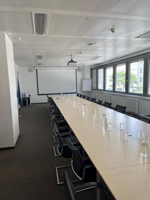 Geräumiger Konferenzraum mit langer Tafel, Beamerleinwand und großen Fenstern für Tageslicht.