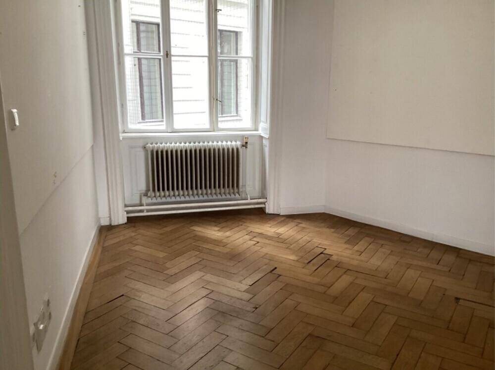 Kleineres Zimmer mit Fischgrätparkett, einem großen Fenster und Heizkörper unter dem Fenster.
