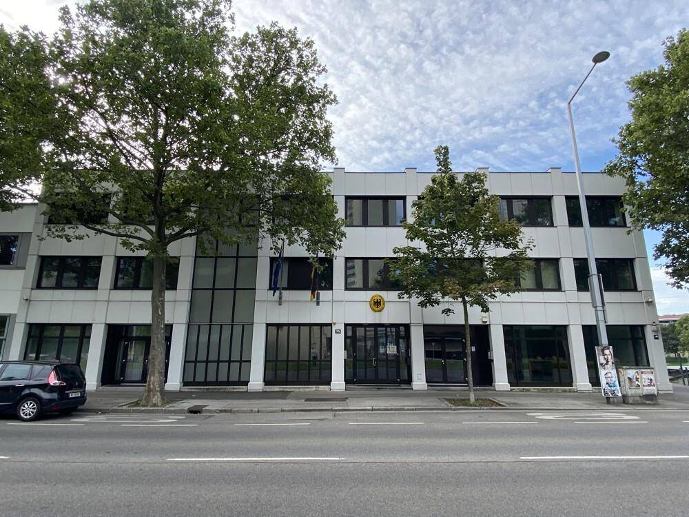 Moderne Gebäudefassade mit großen Fenstern, Bäumen und einer deutschen Flagge an der Vorderseite.