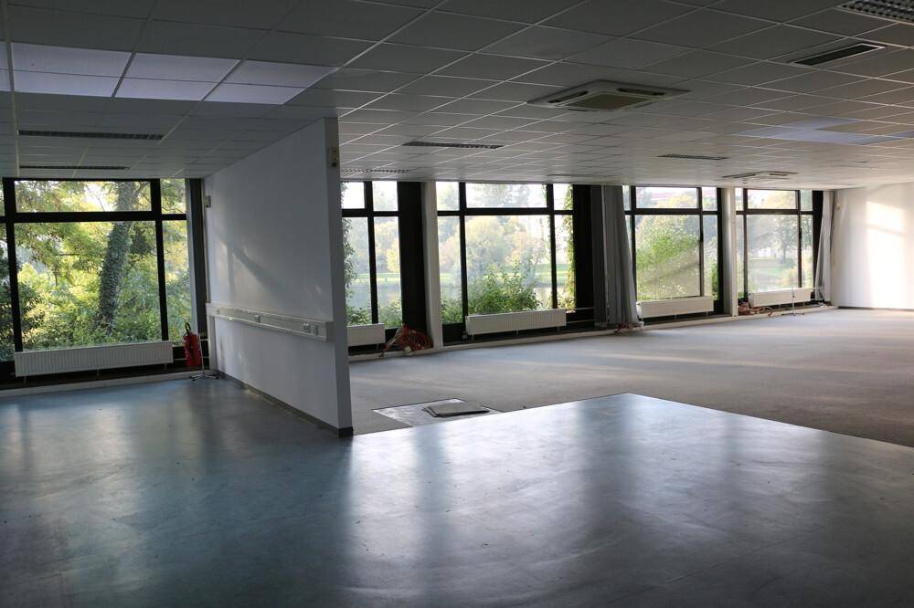 Heller Büroraum mit großen Fenstern, Blick auf Bäume und Wasser, kombiniertem Teppich- und Vinylboden.