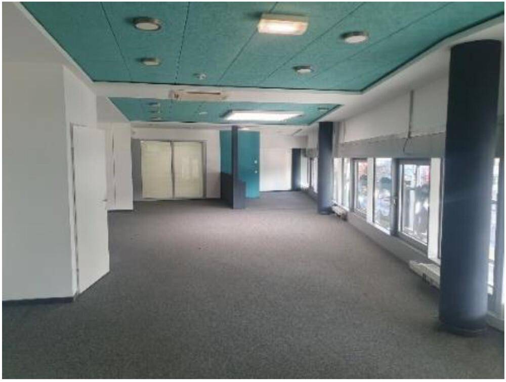 Geräumiger, leerer Büroraum mit großen Fenstern, Säulen und einer markanten grünen Deckenverkleidung.