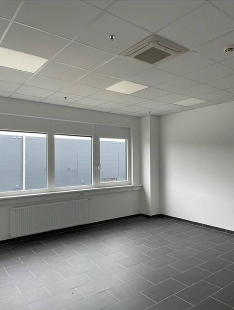 Leerer Büroraum mit grauen Fliesen, weißen Wänden und Fenstern, die Tageslicht hereinlassen.