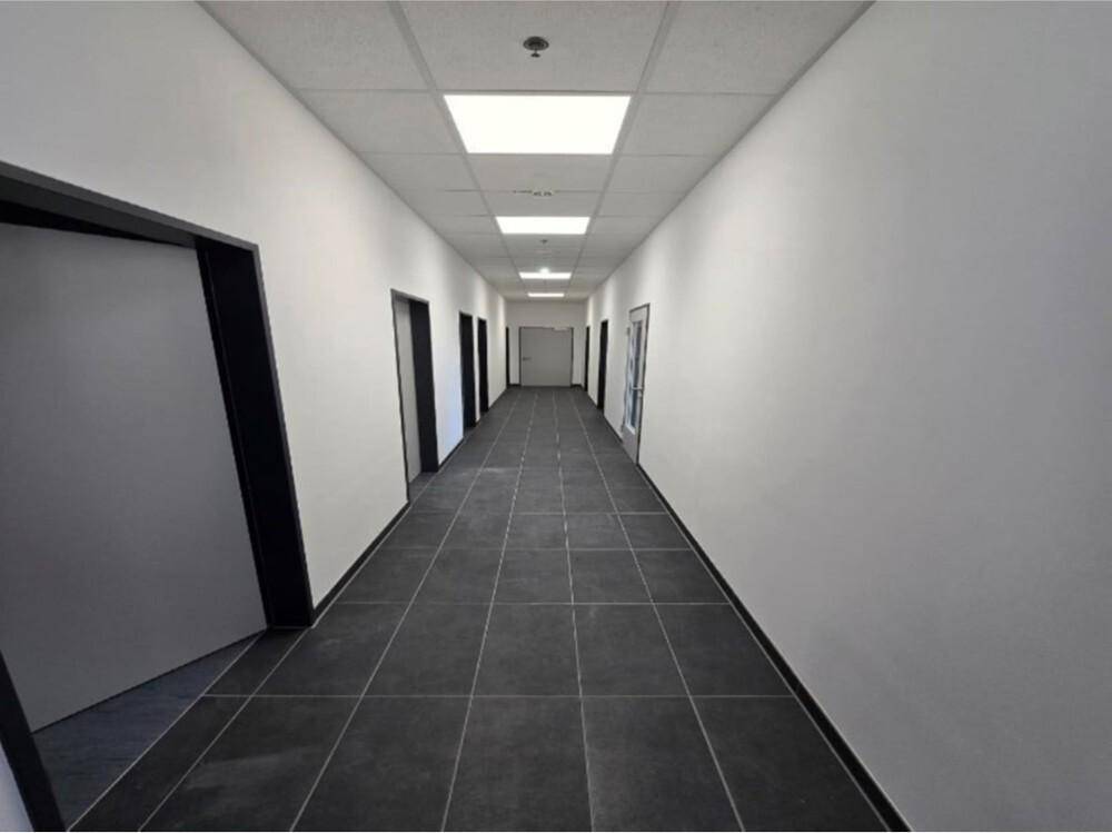 Langer, heller Flur mit dunklen Fliesen, weißen Wänden und Deckenbeleuchtung, der zu mehreren Türen führt.