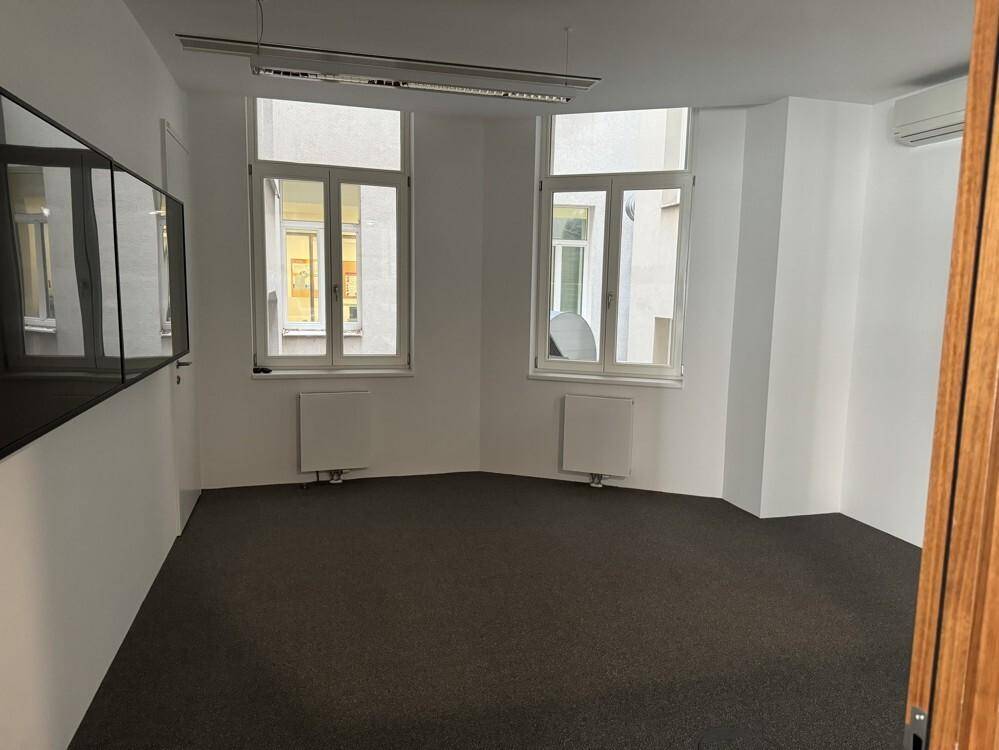 Kleineres Büro mit Teppichboden, zwei Fenstern und Klimaanlage, ideal für Einzelarbeit.
