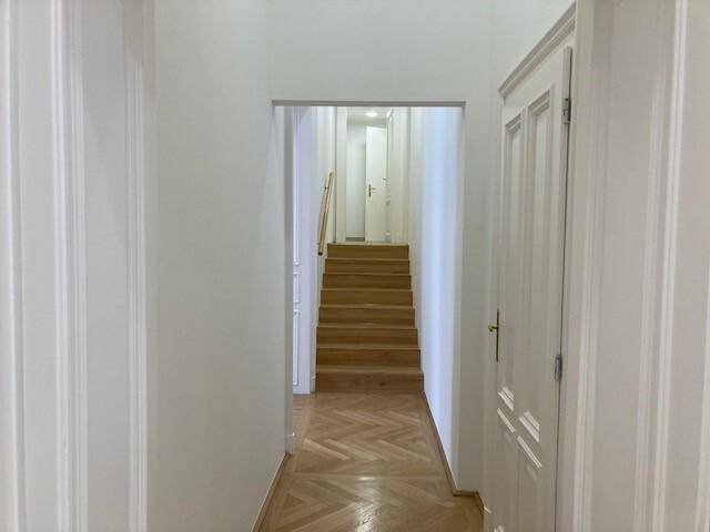 Langer Flur mit Fischgrätparkett, der zu einer Holztreppe führt, umgeben von weißen Wänden und Türen.