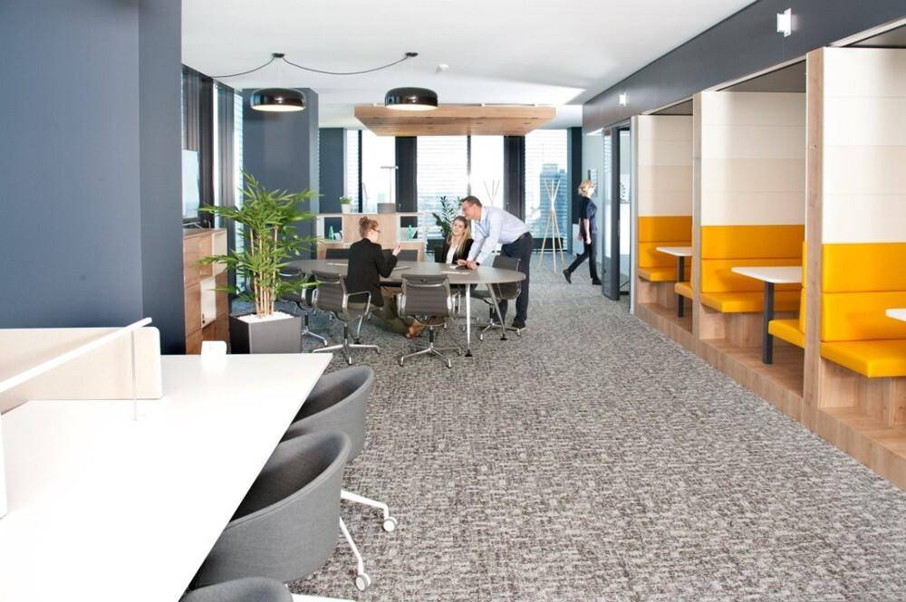 Weitläufiger Großraumbüro-Bereich mit verschiedenen Arbeitszonen, orangefarbenen Sitzkabinen und Besprechungstischen.