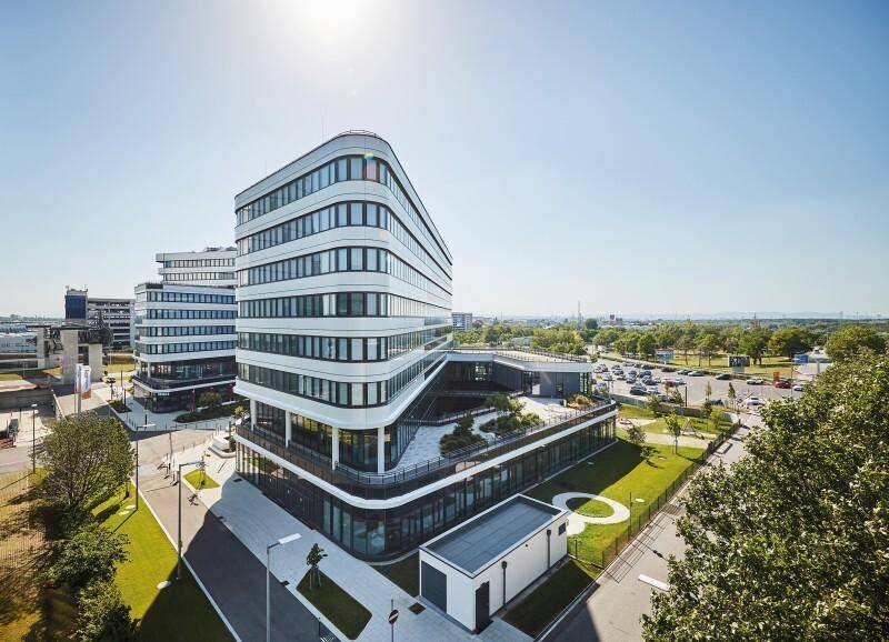Luftaufnahme eines modernen Bürokomplexes mit geschwungener Architektur und begrünten Dachflächen bei Sonnenschein.