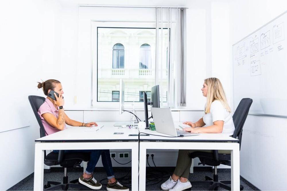 Zwei Personen arbeiten an modernen Schreibtischen in einem hellen Büro mit großem Fenster und Stadtblick.