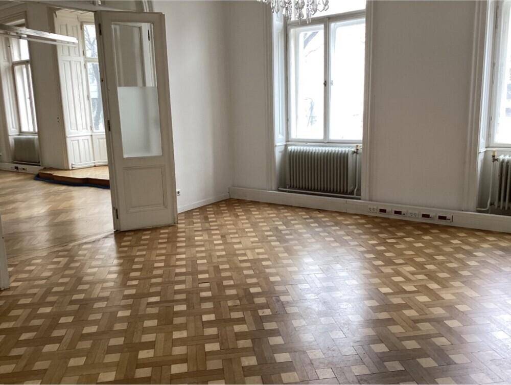 Geräumiges Zimmer mit hellem Parkettboden, hohen Decken und großen Fenstern.
