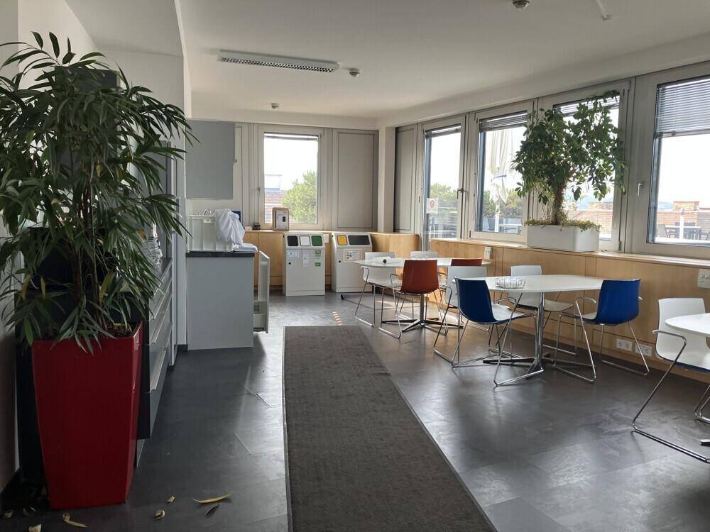 Heller Pausenraum mit Tischen, Stühlen und großen Fenstern, ausgestattet mit Pflanzen und Recyclingstationen.