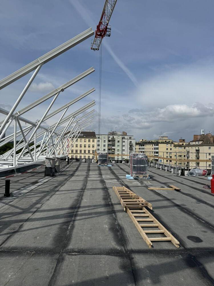 Eine Dachbaustelle mit Metallkonstruktionen und einem Kran, umgeben von städtischen Gebäuden im Hintergrund.