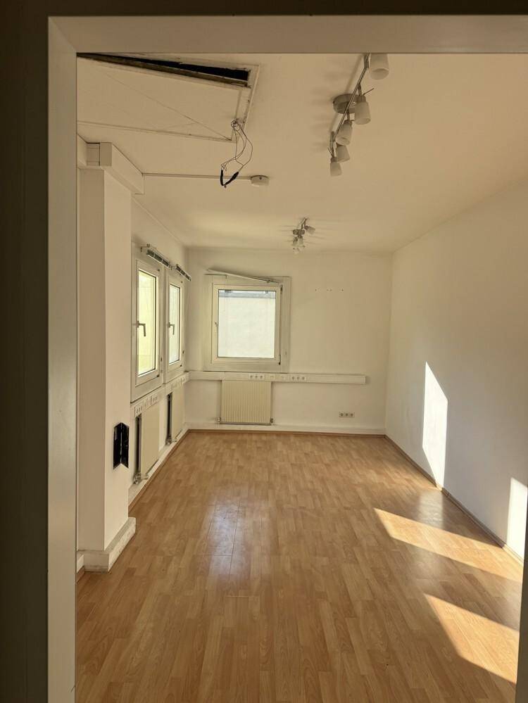 Leerer Büroraum mit Laminatboden, weißen Wänden, großen Fenstern und Deckenstrahlern.
