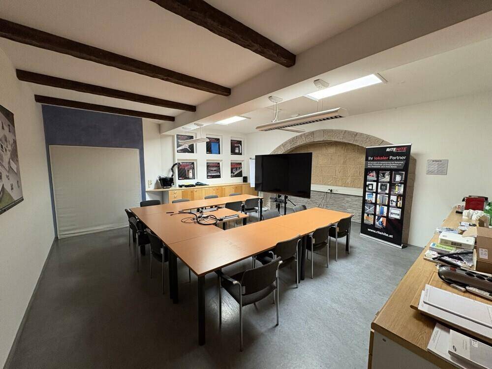 Moderner Konferenzraum mit großem U-förmigen Tisch, Medientechnik und sichtbaren Holzdeckenbalken.