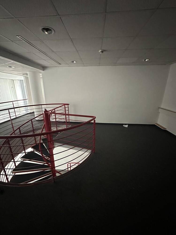 Büroraum mit dunklem Teppichboden, weißen Wänden und einer markanten roten Wendeltreppe, die in eine andere Etage führt.