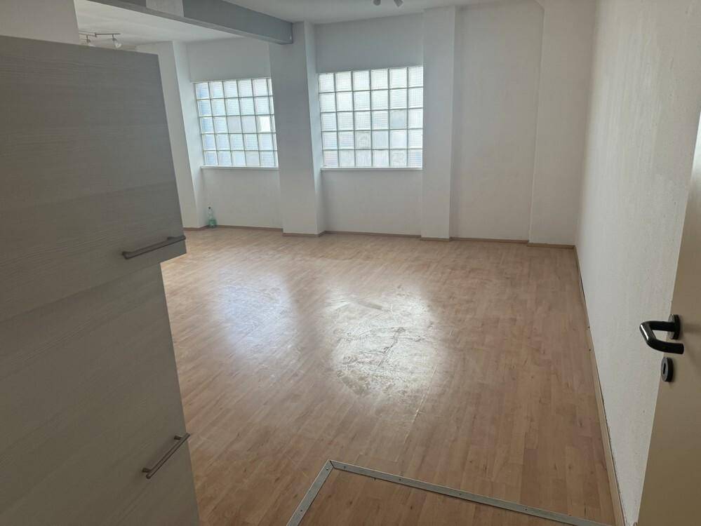 Leerer Raum mit Laminatboden, weißen Wänden und zwei Fenstern aus Glasbausteinen.