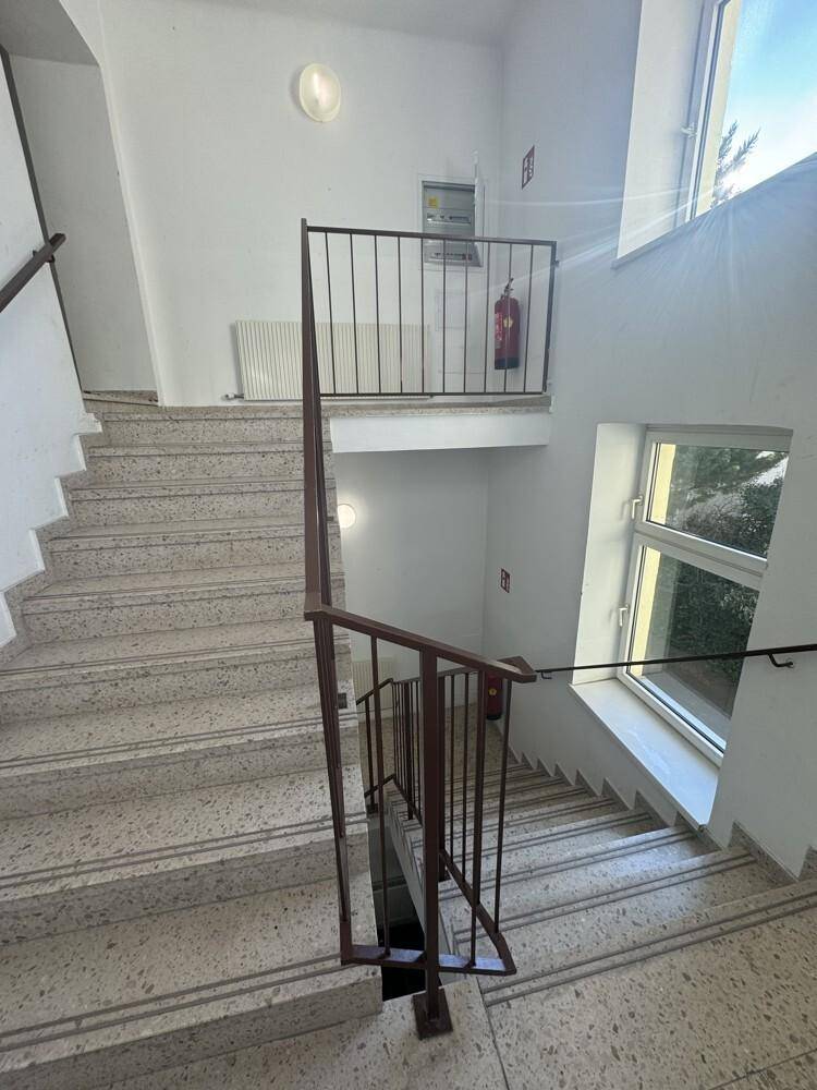 Helles Treppenhaus mit Terrazzostufen, Metallgeländer und großen Fenstern für viel Tageslicht.