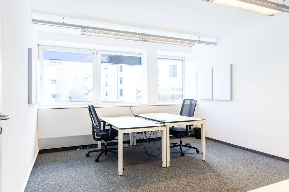 Heller, leerer Büroraum mit zwei Schreibtischen, Bürostühlen und großen Fenstern mit Blick nach draußen.