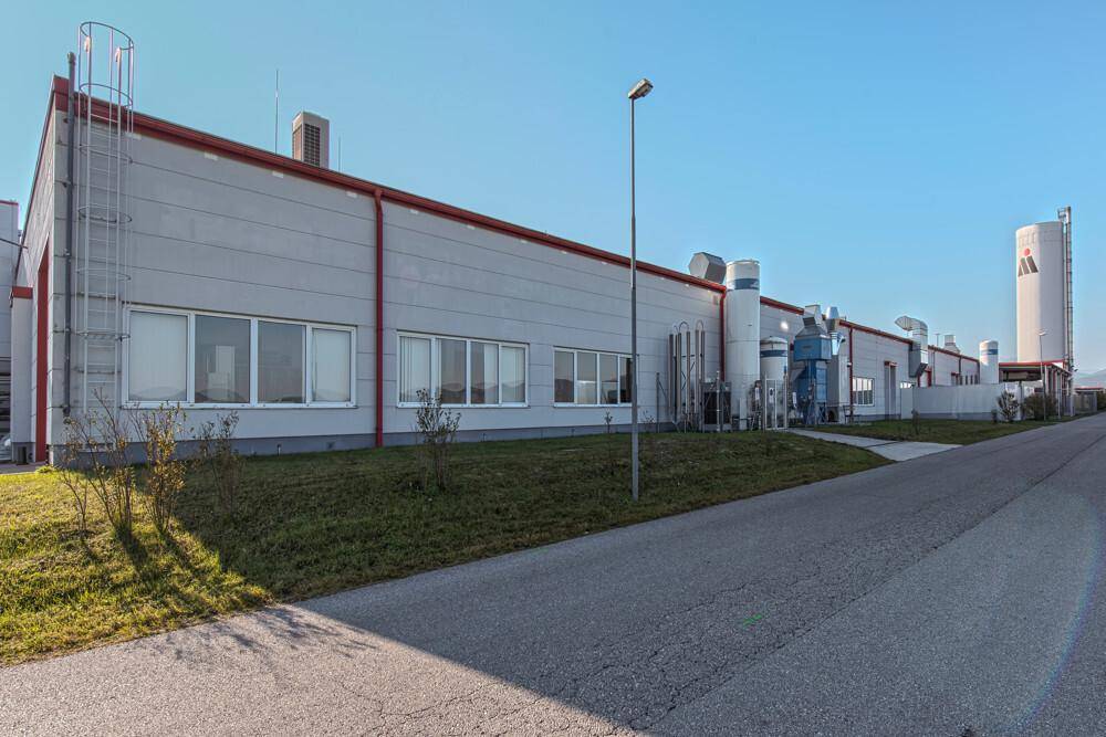 Lange Industriehalle mit technischen Anlagen, Rohren und einem hohen Silo im Hintergrund unter klarem Himmel.