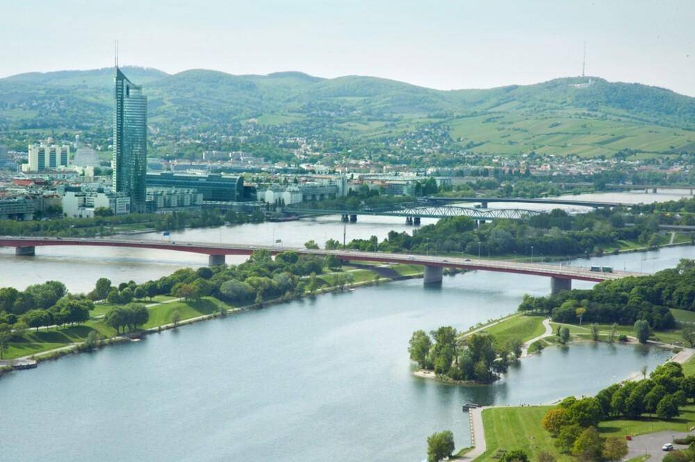 Panoramablick auf eine Stadt mit Fluss, Brücken und einem markanten Hochhaus in der Ferne.