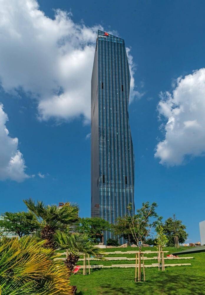 Der schlanke Wolkenkratzer erhebt sich über einer modern gestalteten Parkanlage mit Palmen und terrassierten Grünflächen.