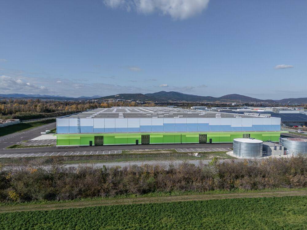 Ansicht der langen, farbigen Fassade des Logistikgebäudes, umgeben von grüner Landschaft und Bergen im Hintergrund.