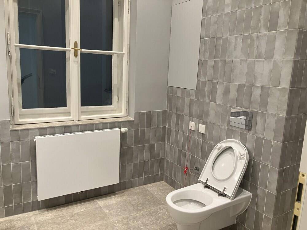 Modernes Badezimmer mit grauen Fliesen, wandhängendem WC und einem großen Fenster für Tageslicht.