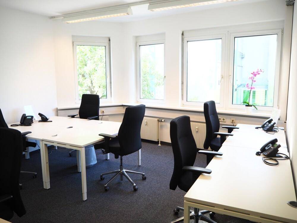 Heller Büroraum mit mehreren Arbeitsplätzen, großen Fenstern und Teppichboden, ideal für ein Team.