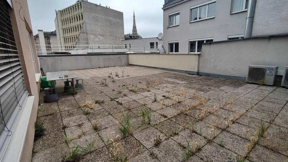 Große Dachterrasse mit Betonplatten und Blick auf die umliegenden Stadtgebäude, benötigt Pflege.