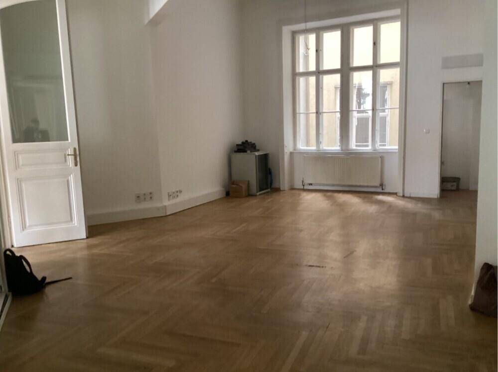 Geräumiges, leeres Zimmer mit Fischgrätparkett, großen Fenstern und viel Tageslicht.
