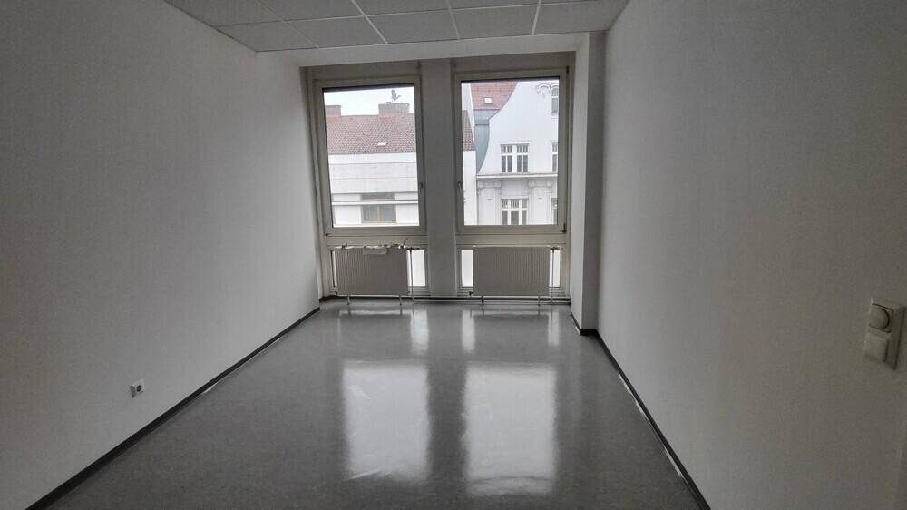 Kleiner, heller Büroraum mit grauem Boden und zwei großen Fenstern, die Tageslicht hereinlassen.