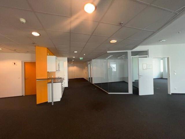 Offener Bürobereich mit Teppichboden, Glaswänden und einer modernen Küchenzeile mit orangefarbenem Akzent.