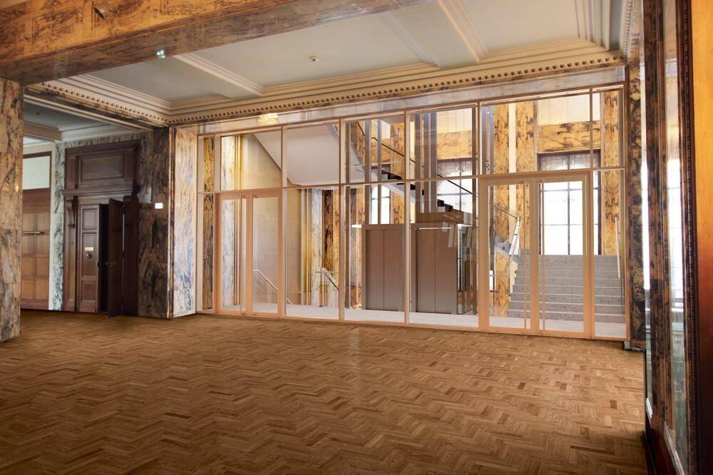 Eleganter Eingangsbereich mit glänzendem Fischgrätparkett, Marmorverkleidungen und einer modernen Glasfront zum Treppenhaus und Aufzug.