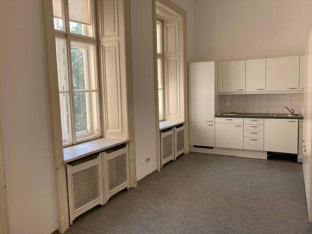Geräumiger Raum mit weißer Einbauküche und zwei großen Fenstern, die viel Licht hereinlassen.
